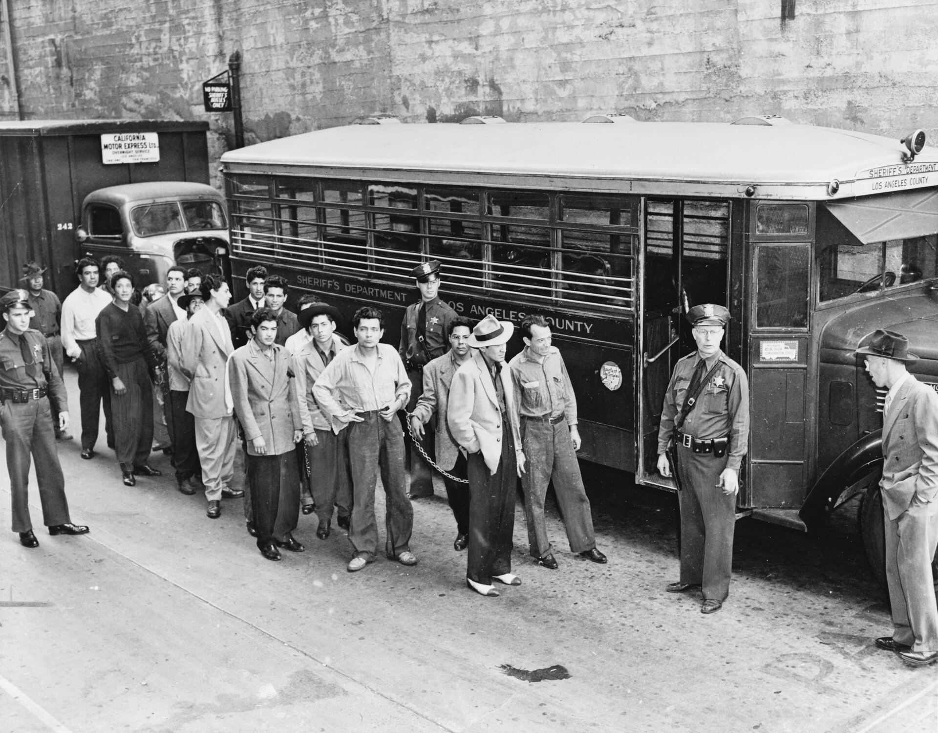 Young Mexican American men in zoot suits lined up beside a Los Angeles County Sheriff bus, guarded by deputies, in June 1943